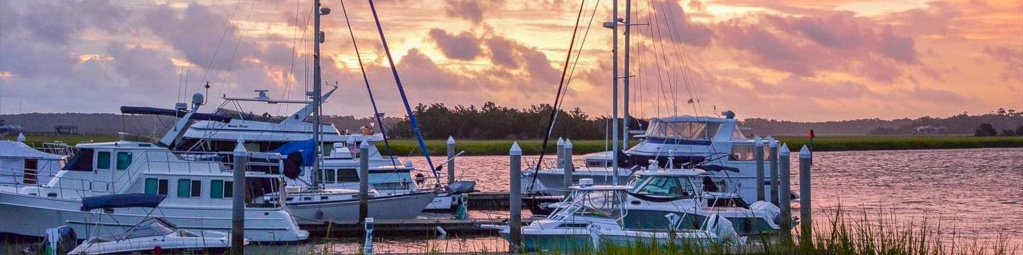 A serene marina scene at sunset with sailboats and yachts docked, vibrant sky hues reflecting on calm water.