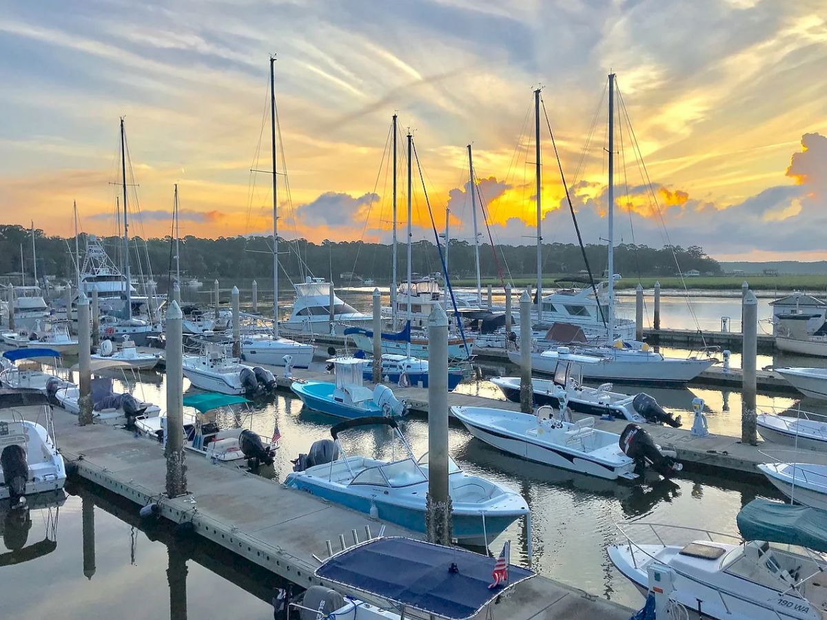 The image shows a marina with multiple boats and yachts docked, set against a beautiful sunrise or sunset sky with dramatic clouds and reflections in the water.