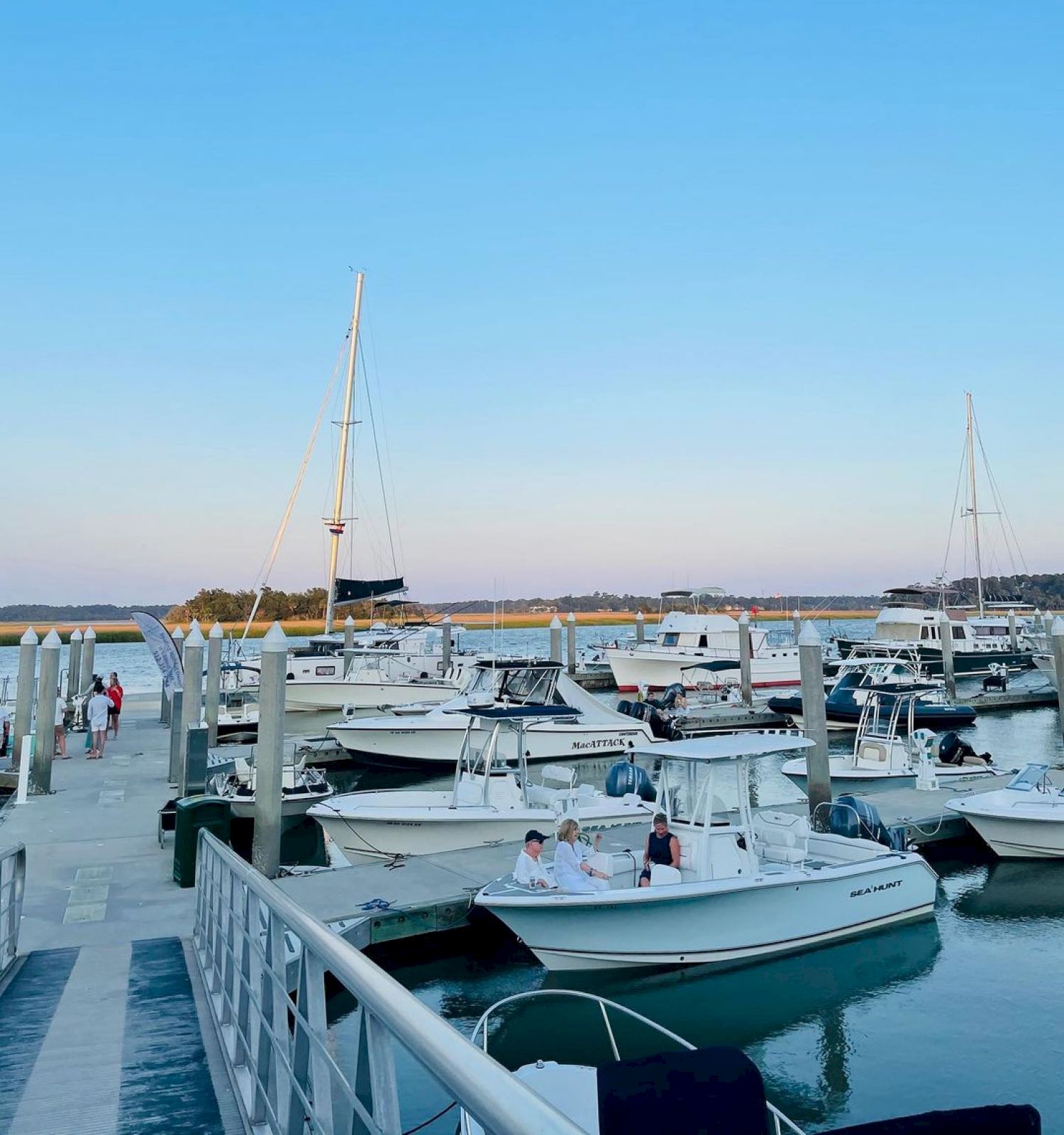 The image shows a marina with various boats docked, a clear blue sky, calm water, and a few people walking on the dock.