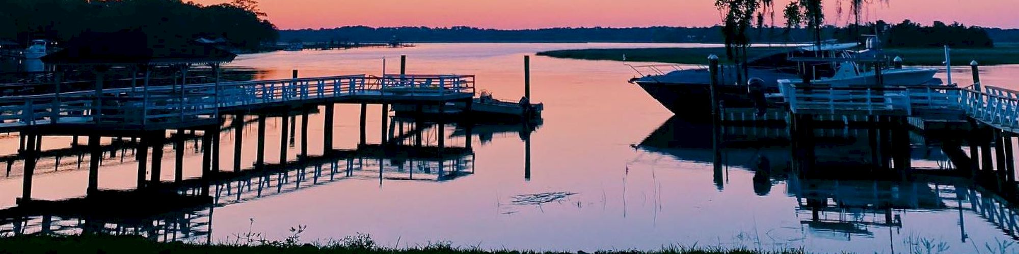 A serene waterfront at sunset with boats docked at piers, reflecting on the calm water, and trees hanging over the scene from above, ending the sentence.