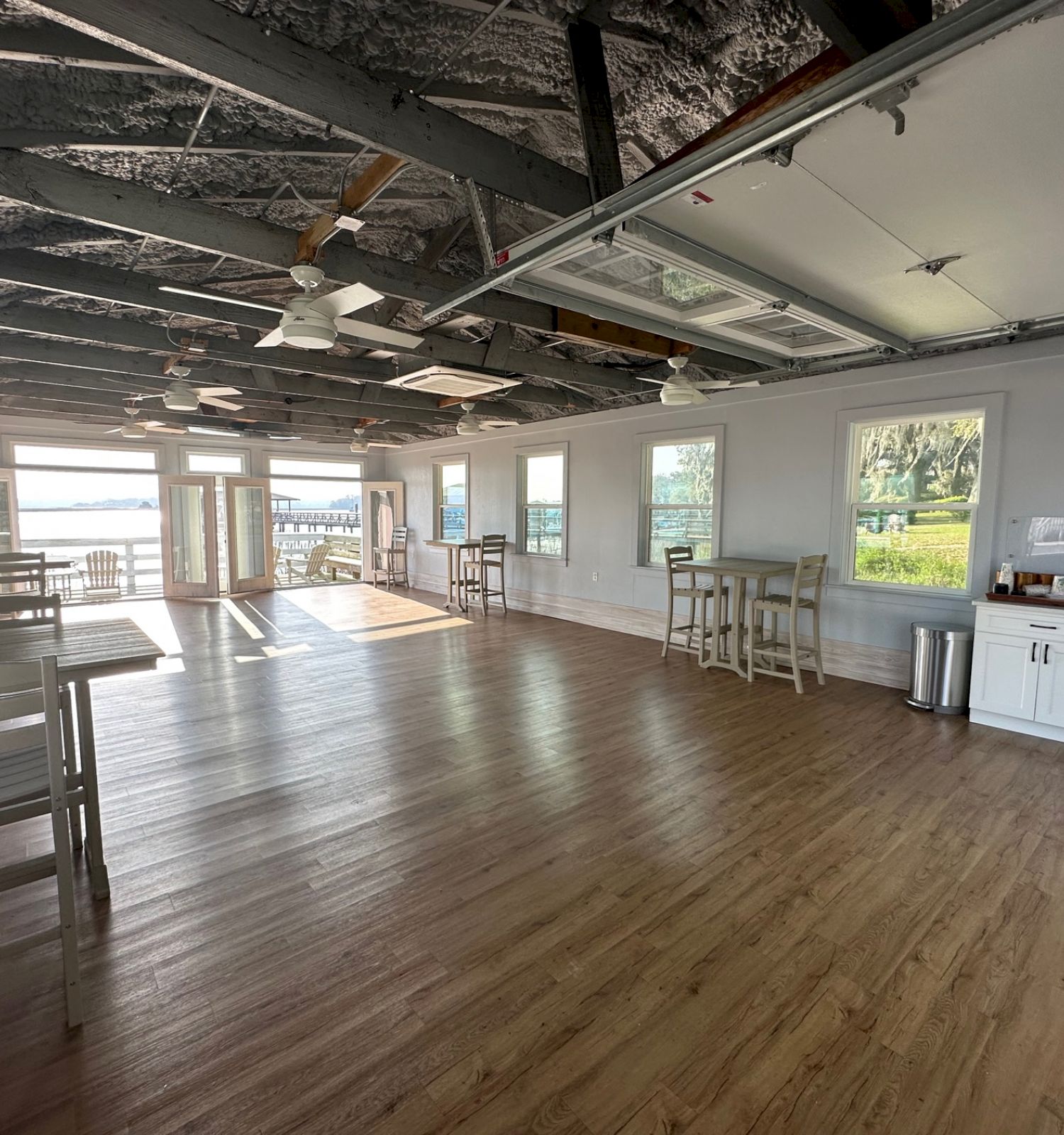 An empty, spacious room with wooden floors, tables, chairs, large windows, ceiling fans, and a coffee station, overlooking a scenic outdoor view.