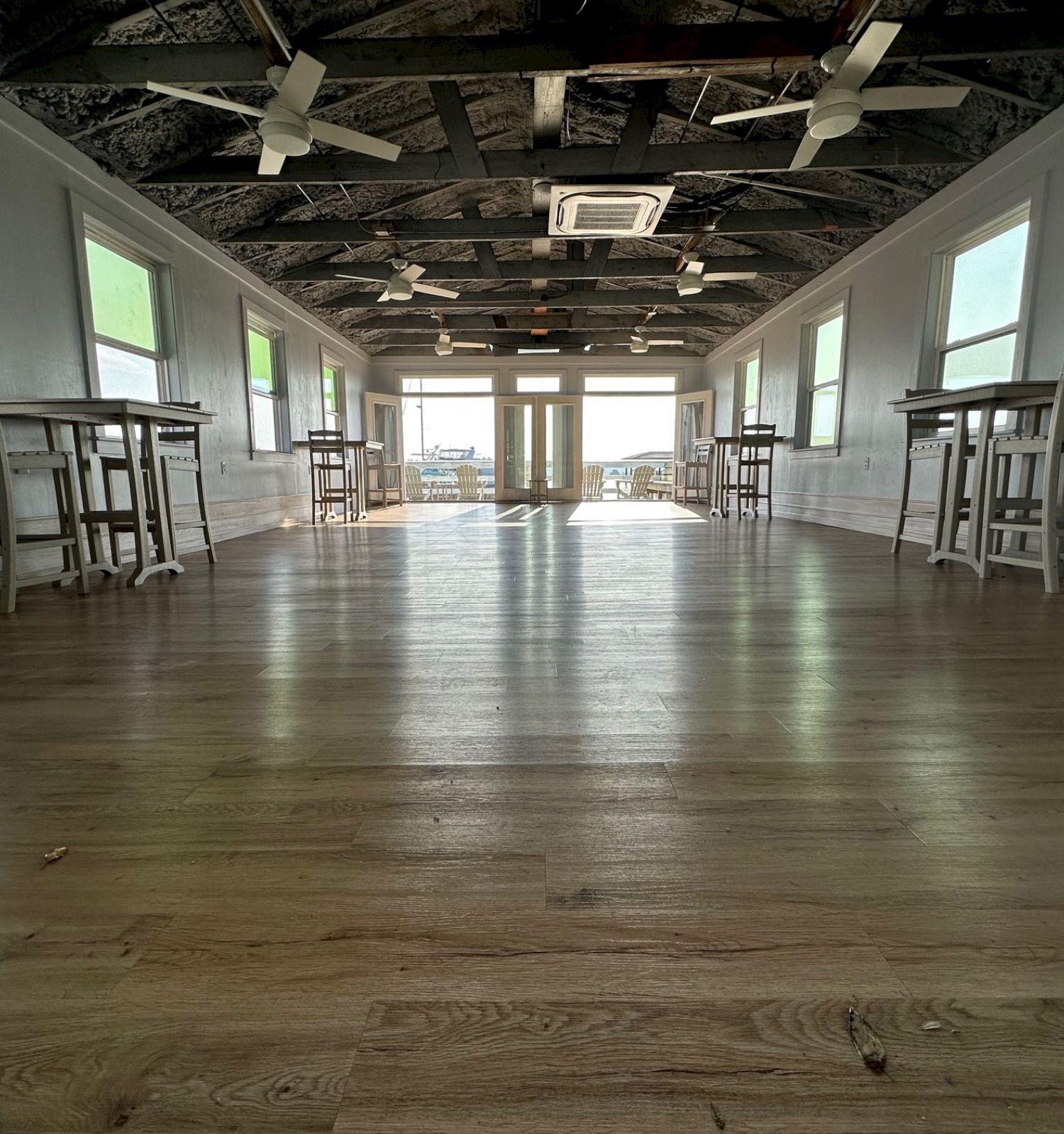 A spacious hall with wood flooring, high ceiling with fans, large windows, and minimal wooden tables and chairs against the walls, leading to glass doors.