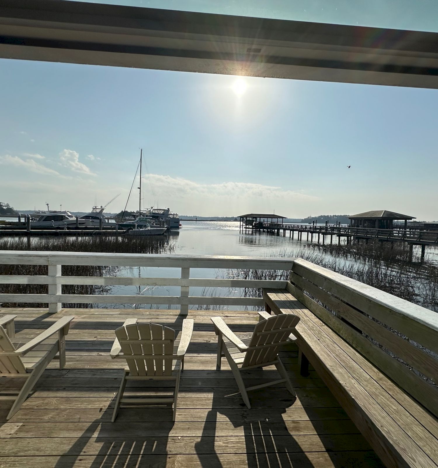 A peaceful waterfront view with a wooden deck, three chairs, and boats docked under the sun’s warm glow, framed by a window.