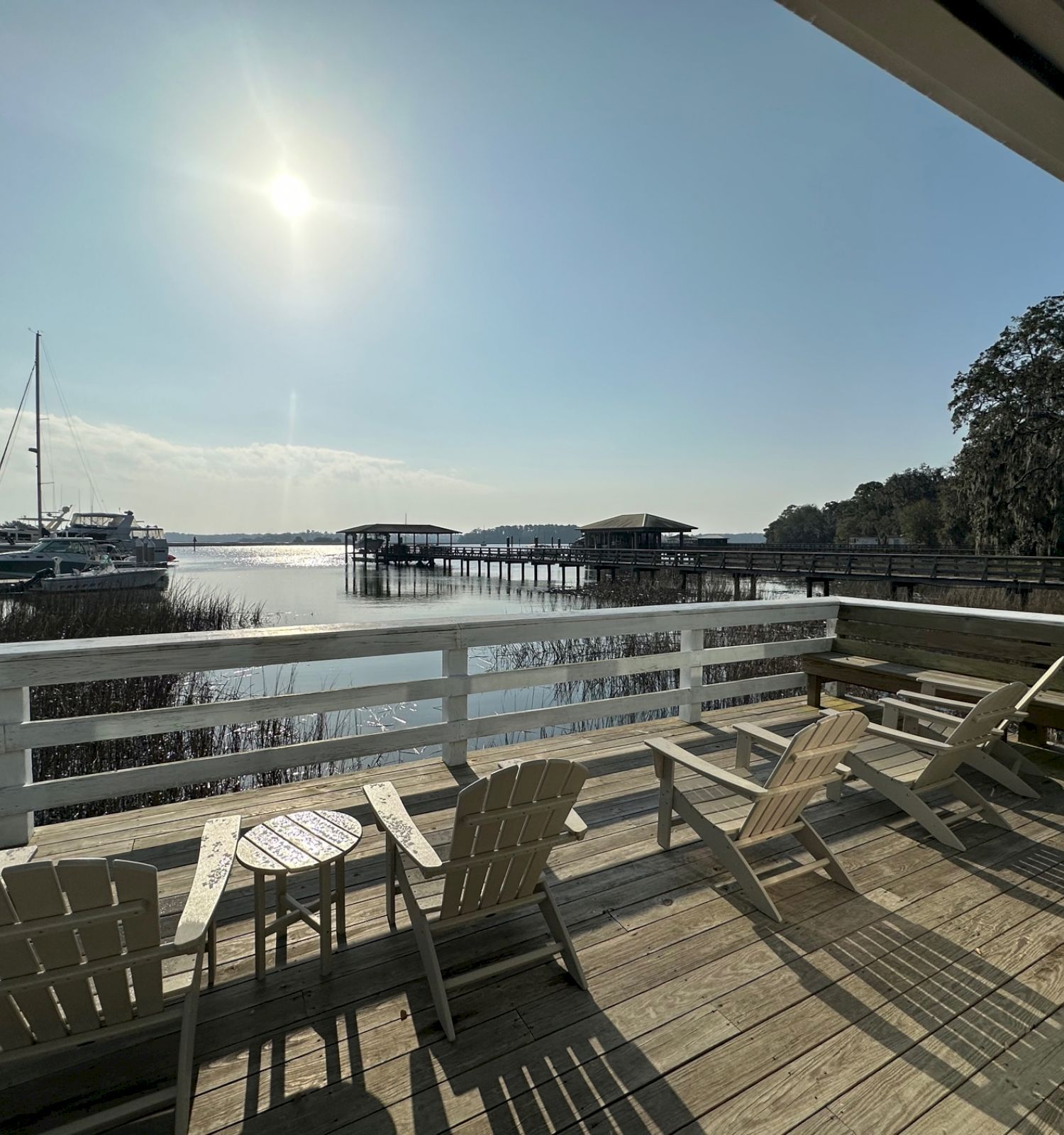 The image shows a sunlit deck with Adirondack chairs, overlooking boats on the water and a wooden dock, framed by trees under a clear sky.