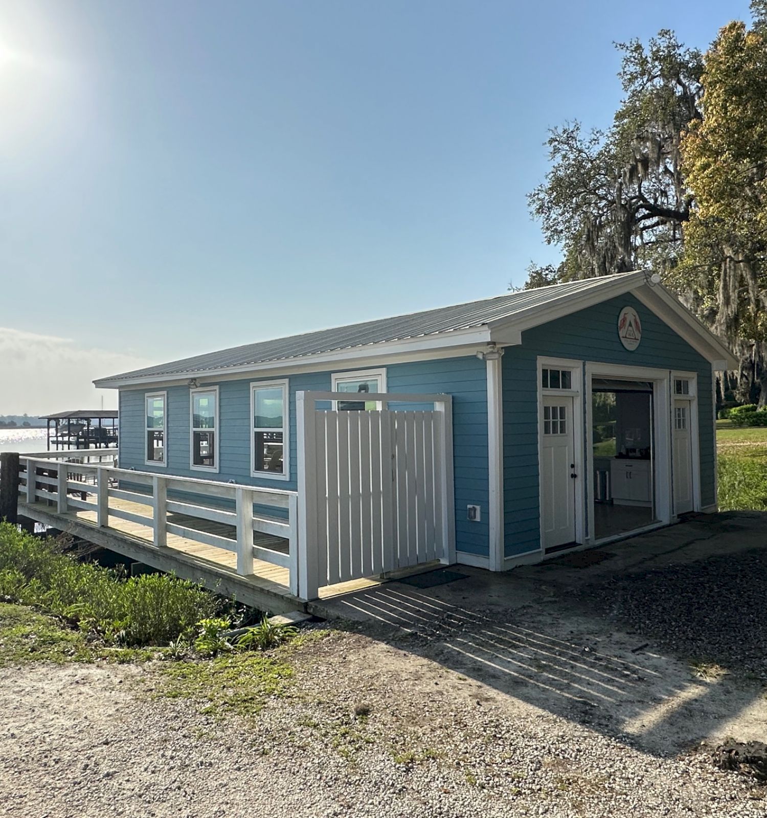 This image shows a light blue cabin with white trim, located near a body of water, with a pier and greenery around it, under a bright sky.