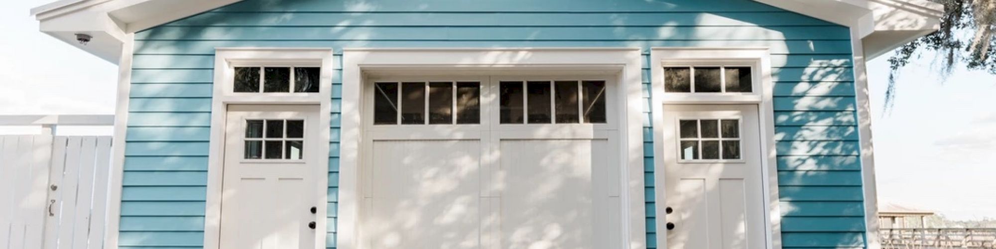 A blue garage with white trim features two white side doors and a central, larger door, alongside a gravel driveway and some trees in the background.