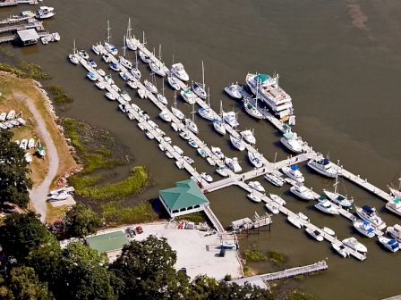 An aerial view of a marina with multiple boats docked at piers, surrounded by water and nearby buildings and greenery, ending the sentence.