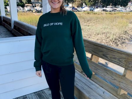 A person stands smiling on a wooden deck by the water, wearing a green "Isle of Hope" sweatshirt, black leggings, and sneakers, with trees in the background.