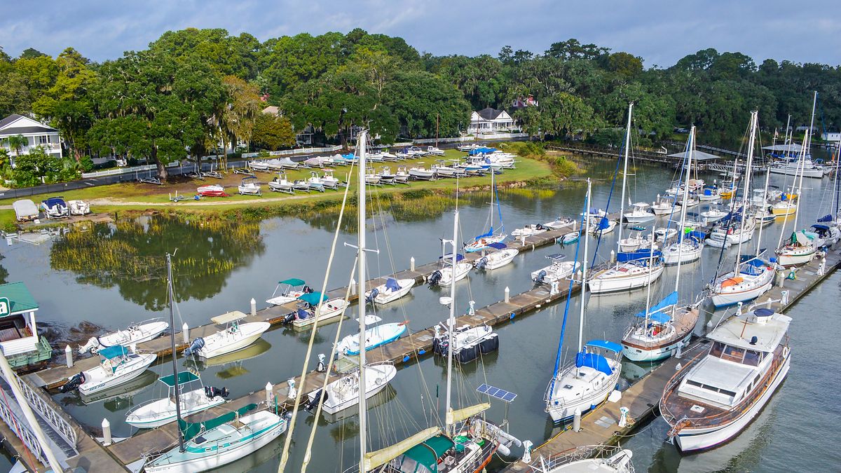 A marina with numerous sailboats and yachts docked, surrounded by lush greenery and residential houses in the background, partially cloudy sky.