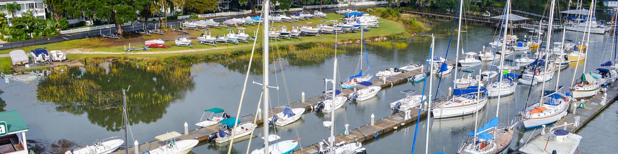 A marina with numerous sailboats and yachts docked, surrounded by lush greenery and residential houses in the background, partially cloudy sky.