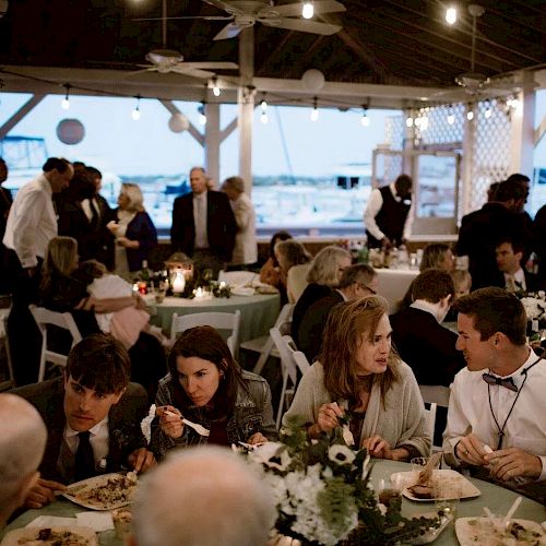 People are dining and socializing under a pavilion with string lights, suggesting a gathering or celebration by the water.