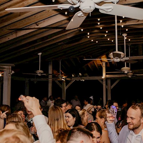 People are dancing and celebrating under string lights and ceiling fans in a wooden structure.