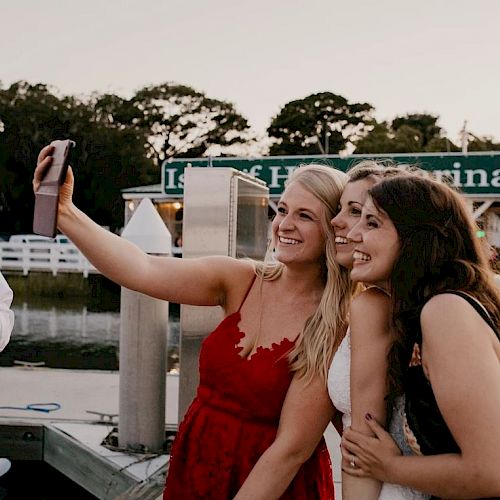 Three people are taking a group selfie near a marina at sunset. A green sign in the background reads 