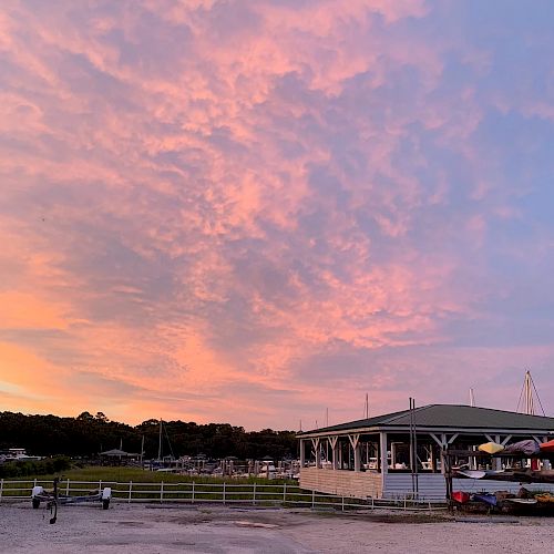 The image shows a picturesque sunset with a stunning pink and orange sky above a waterfront area. There's a pavilion and a dock with boats.