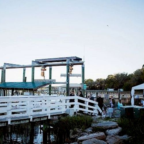 A waterfront scene with structures on a pier, white fencing, a tent, and people milling about near the water, with trees in the background.