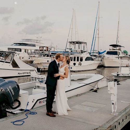 A couple in formal attire embraces on a dock with multiple boats in the background, under a partially cloudy sky with a visible moon.