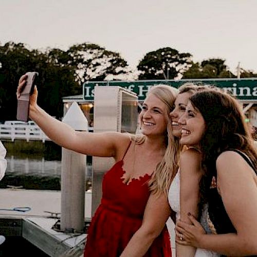 Three people are taking a selfie near a marina. Two women are smiling and posing while the man is adjusting his tie to the left of the frame.