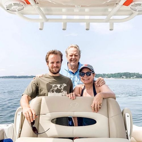 Three people are on a boat, posing for a photo with water and distant trees in the background. They're smiling and enjoying a sunny day.