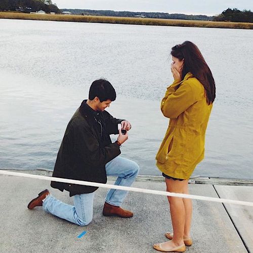 A person is kneeling and holding up a ring in front of another person who appears emotional, near a waterfront.