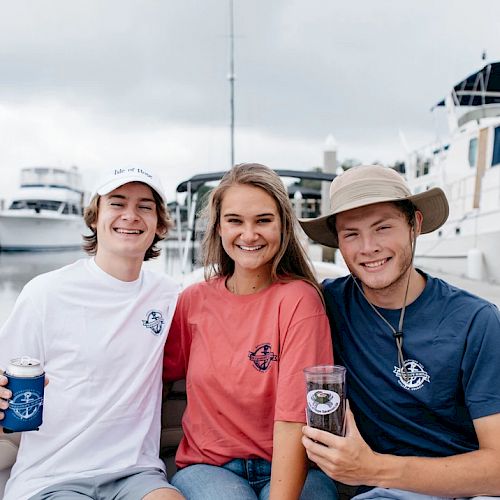 Three smiling people sitting on a boat, holding drinks, with a marina and yachts in the background. They wear casual boating attire.