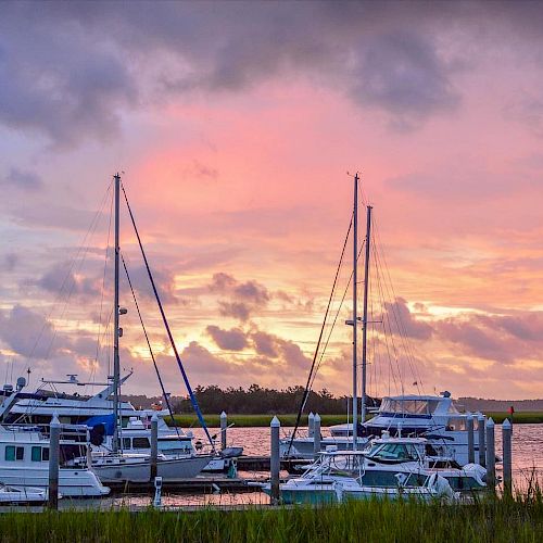 Several boats are docked at a marina with a scenic sunset and cloudy sky in the background.