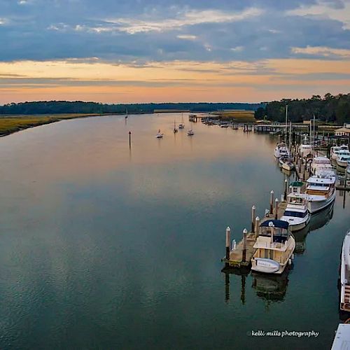 A serene marina with numerous docked boats during sunset, reflecting the sky and offering a tranquil waterfront scene. The photo is by 'bella-mills photography'.