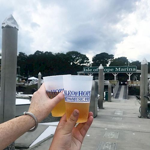 Two people are holding drinks in plastic cups at Isle of Hope Marina, with docks and boats visible in the background on a cloudy day.