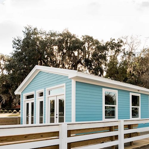 A small, light blue building with white trim, surrounded by trees and a white fence, under a partially cloudy sky.