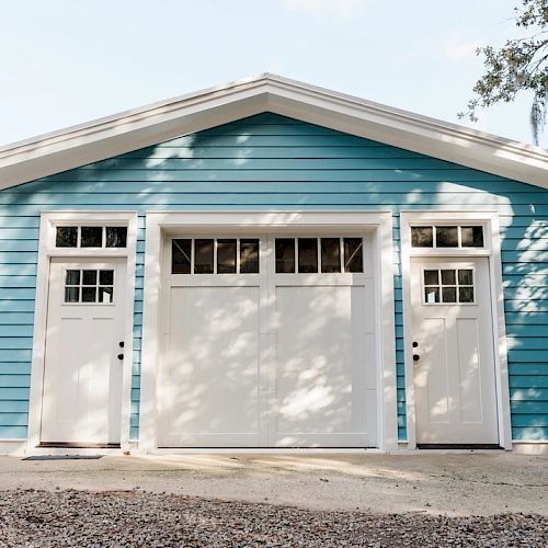 A blue garage with white trims, featuring a large central door and two smaller doors on each side. Trees and natural surroundings are visible.