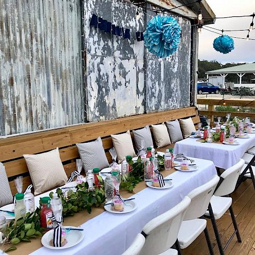 A decorated outdoor dining setup with white tablecloths, arranged plates, greenery, blue decorative balls, and string lights, next to a rustic wall.