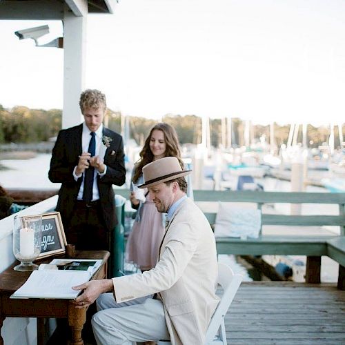 Three individuals, one playing piano and two watching near docks, with boats in the background. A small sign with 