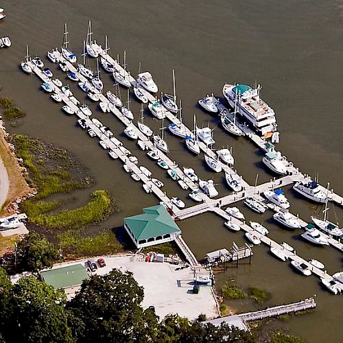 The image shows an aerial view of a marina with multiple boats docked, a small building with a green roof, and surrounding greenery and structures.