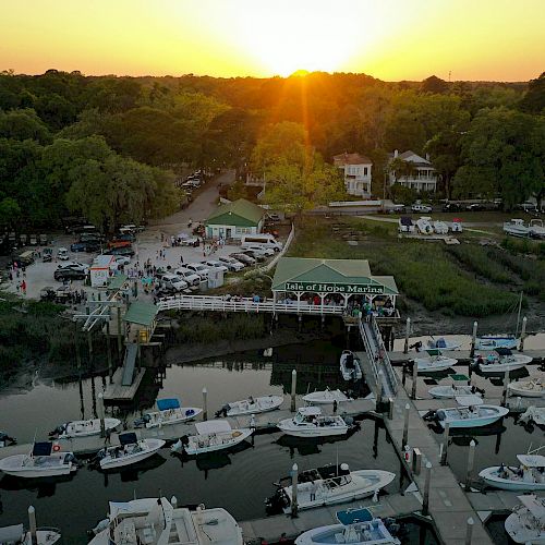 A picturesque marina at sunset with boats docked, a wooden pier, nearby buildings, and a vibrant orange sky over surrounding greenery ending the sentence.