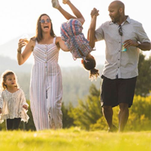 A family enjoys a sunny day outdoors. The father is playfully swinging a child upside down while the mother and another child walk beside them, all smiling.