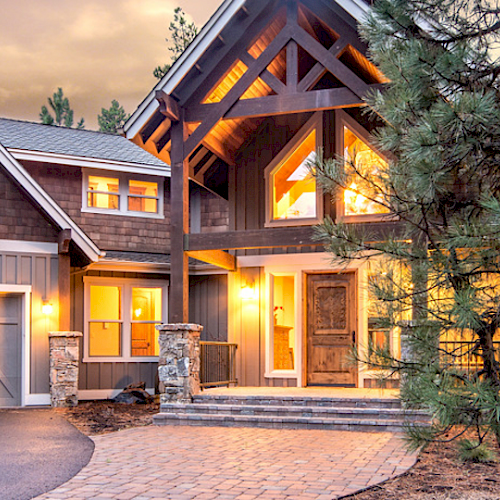 This image shows a two-story modern house with large windows and a three-car garage, surrounded by trees and illuminated warmly.