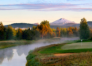 A serene golf course with a green and flag beside a reflective water body, surrounded by misty trees and mountains, with a snow-capped peak in the background.