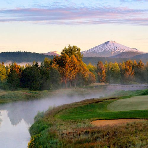 A serene golf course with a green and flag beside a reflective water body, surrounded by misty trees and mountains, with a snow-capped peak in the background.