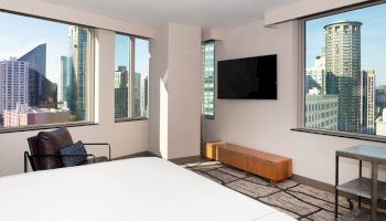 A modern hotel room with large windows showcasing a cityscape, a wall-mounted TV, a chair, and a minimalist decor.