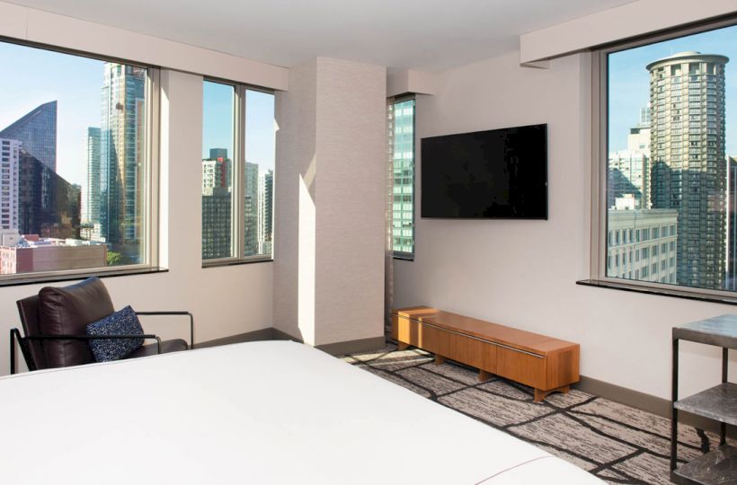 A modern hotel room with large windows showcasing a cityscape, a wall-mounted TV, a chair, and a minimalist decor.