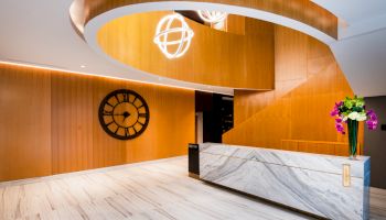 A modern lobby with wooden accents, a large clock on the wall, marble reception desk, spiral staircase, and pendant lights.