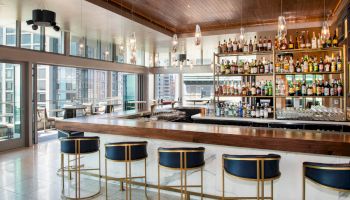 A modern bar with a wooden countertop, blue and gold stools, and a wall of liquor bottles. Large windows offer a view of the city skyline.