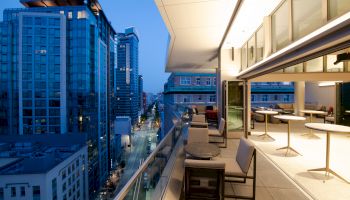 A modern balcony view of a city at dusk, featuring high-rise buildings, outdoor seating, and a well-lit interior space through large windows.