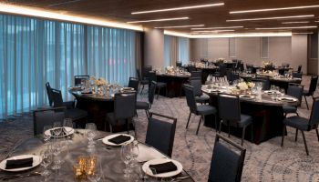 A banquet hall is set up with round tables, chairs, and table settings, including folded napkins and glassware, under warm ceiling lights.