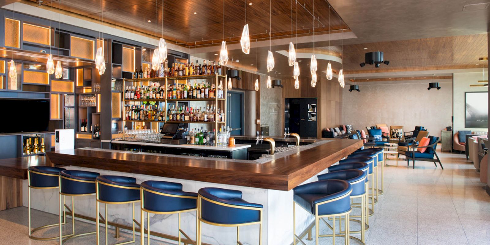 This image shows a modern bar with a wooden counter surrounded by blue bar stools. It has a large selection of bottles and hanging light fixtures.