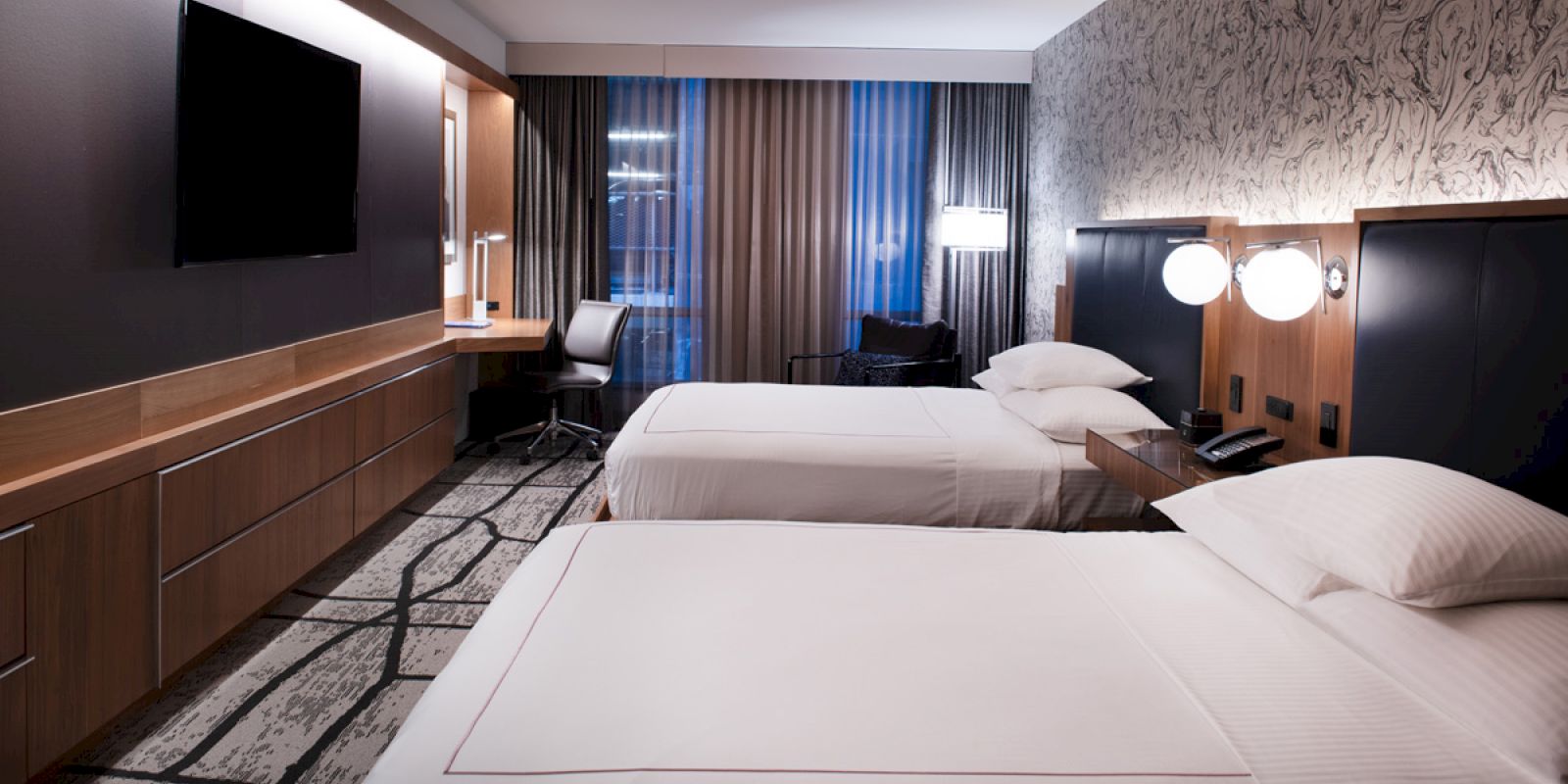 A modern hotel room features two neatly made beds, a wall-mounted TV, a desk with a chair, and contemporary decor with stylish lighting.