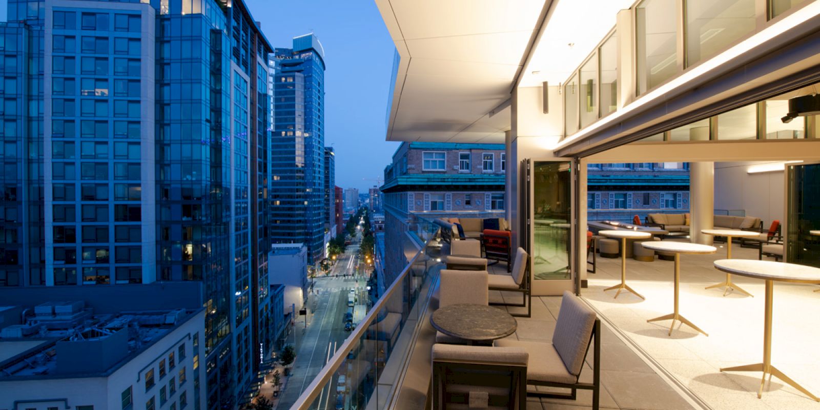 A modern cityscape view at dusk from a high-rise balcony furnished with tables and chairs, overlooking illuminated skyscrapers and a busy street.