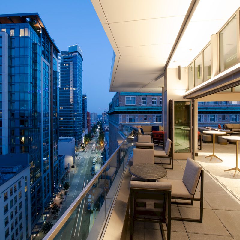 A modern balcony with tables and chairs overlooks a busy urban street lined with tall buildings at dusk.