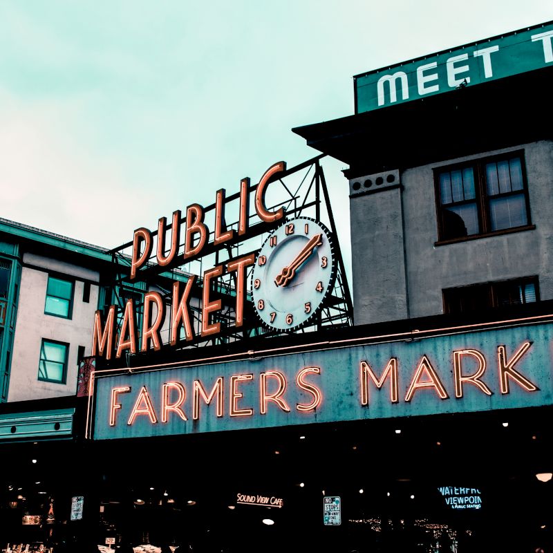 This image shows a busy market area with vibrant signs that read "Public Market" and "Farmers Market." There are surrounding buildings and a clock.