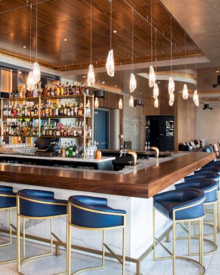 This image shows a modern bar with a wooden counter, surrounded by blue and gold stools, and a well-stocked back bar, under stylish pendant lights.