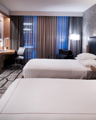 A modern hotel room with two beds, wall-mounted TV, desk, and chair by the window. The room features contemporary décor and soft lighting.